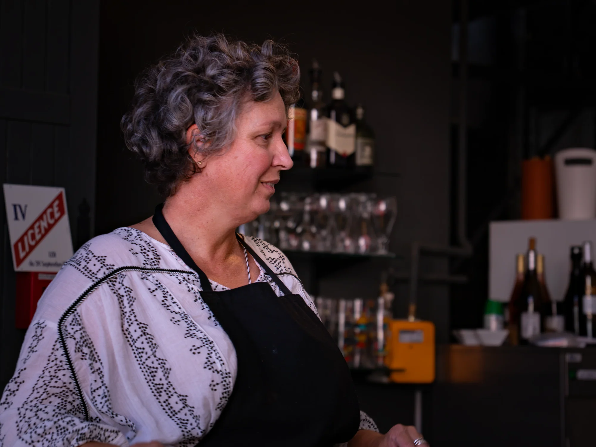 Marie-Gabrielle de profil, en tablier noir, derrière le comptoir de la rôtisserie